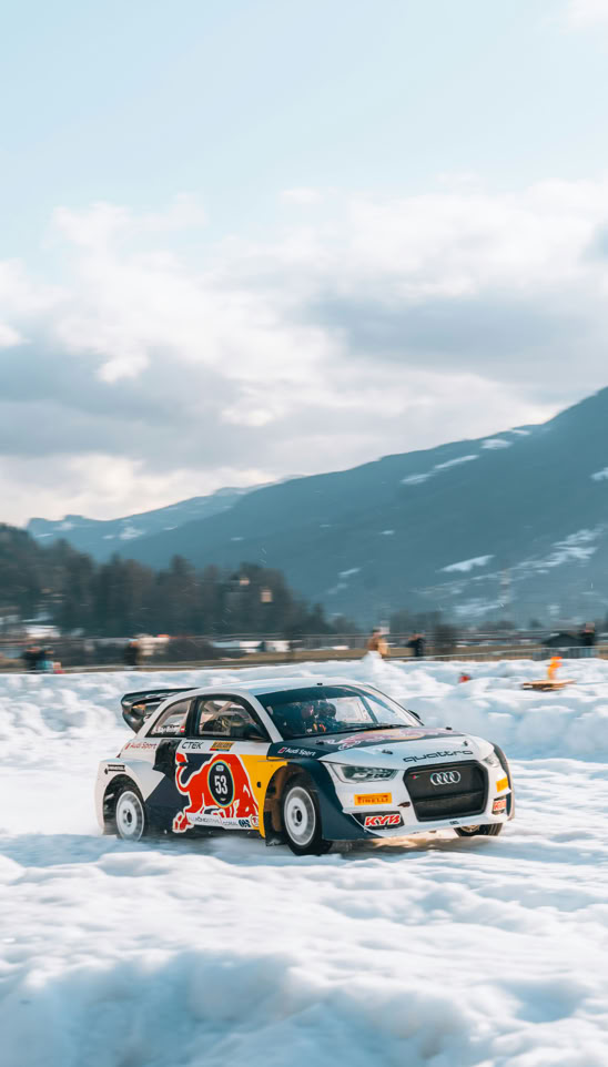 A rally car with colorful graphics driving on a snowy landscape, with mountains and a cloudy sky in the background.